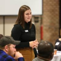 A student smiles standing at a round table of attendees